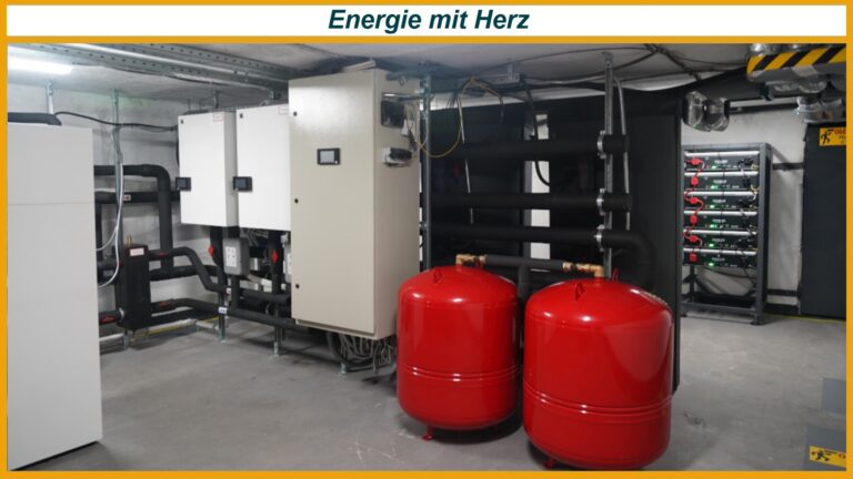 Zwei rote Ausdehnungsbehälter vor Rohrleitungen, Steuerkästen und Batterieregalen in Technikraum; Banner 'Energie mit Herz' oben.